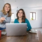 Madre feliz trabajando en laptop desde su hogar, mientras su hijo juega, conciliando vida familiar y negocios desde casa para mujeres con hijos.